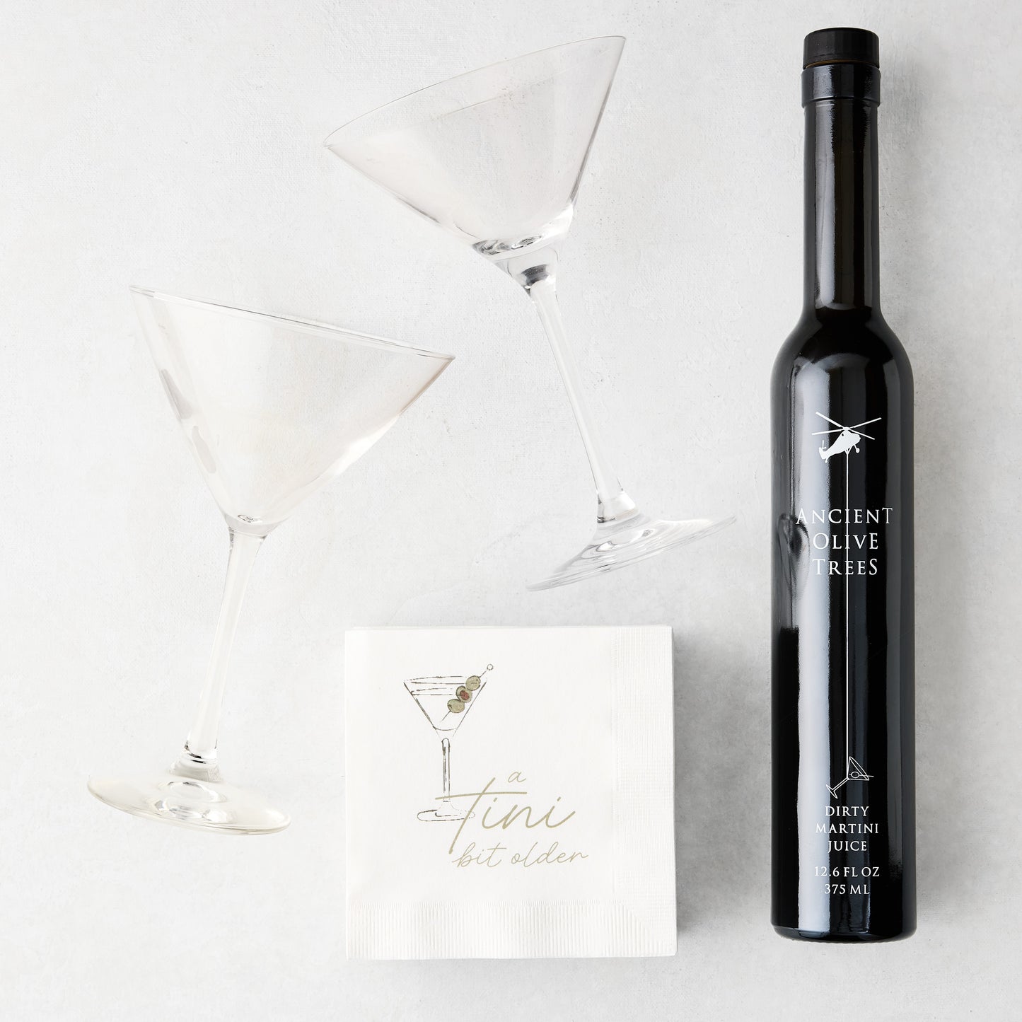 Two clear martini glasses, a black bottle of Ancient Olive Trees Dirty Martini Juice, and white "a tini bit older" cocktail napkins.