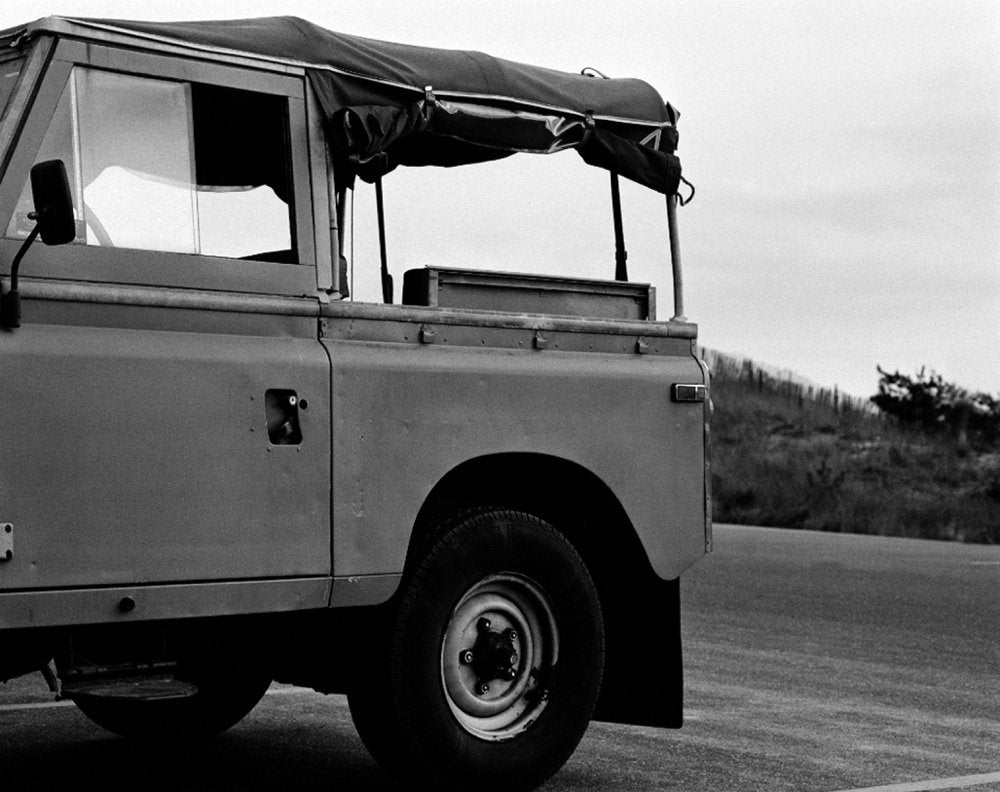 Black and white photograph of a vintage surf truck with a soft top, parked on a road by sand dunes.