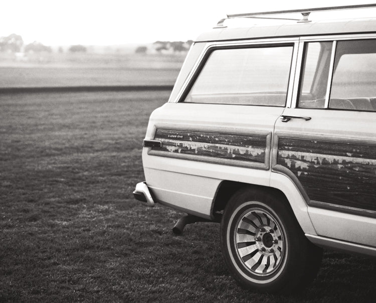 Black and white photograph of a vintage white Wagoneer with wood paneling parked in a grassy field.