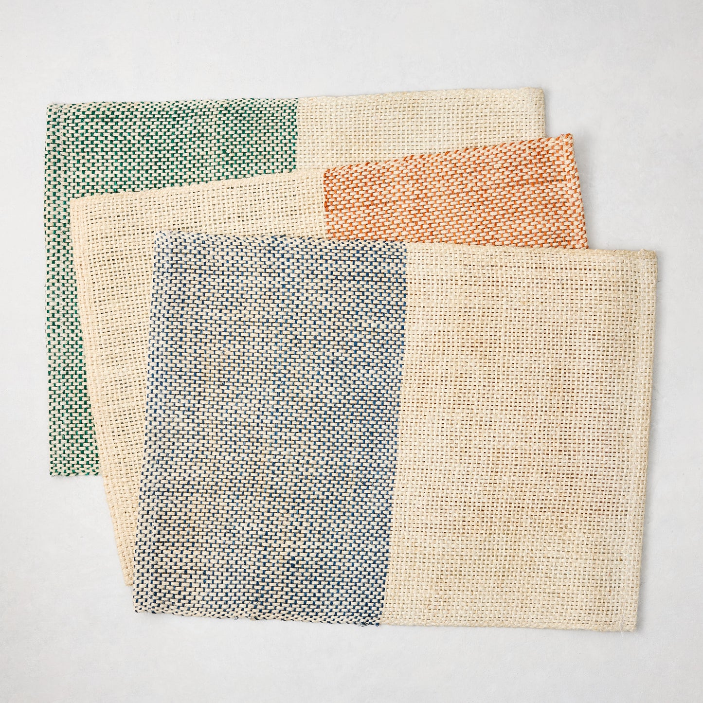 Three woven abaca and jute placemats: blue, orange, and green half and half patterns with natural beige.