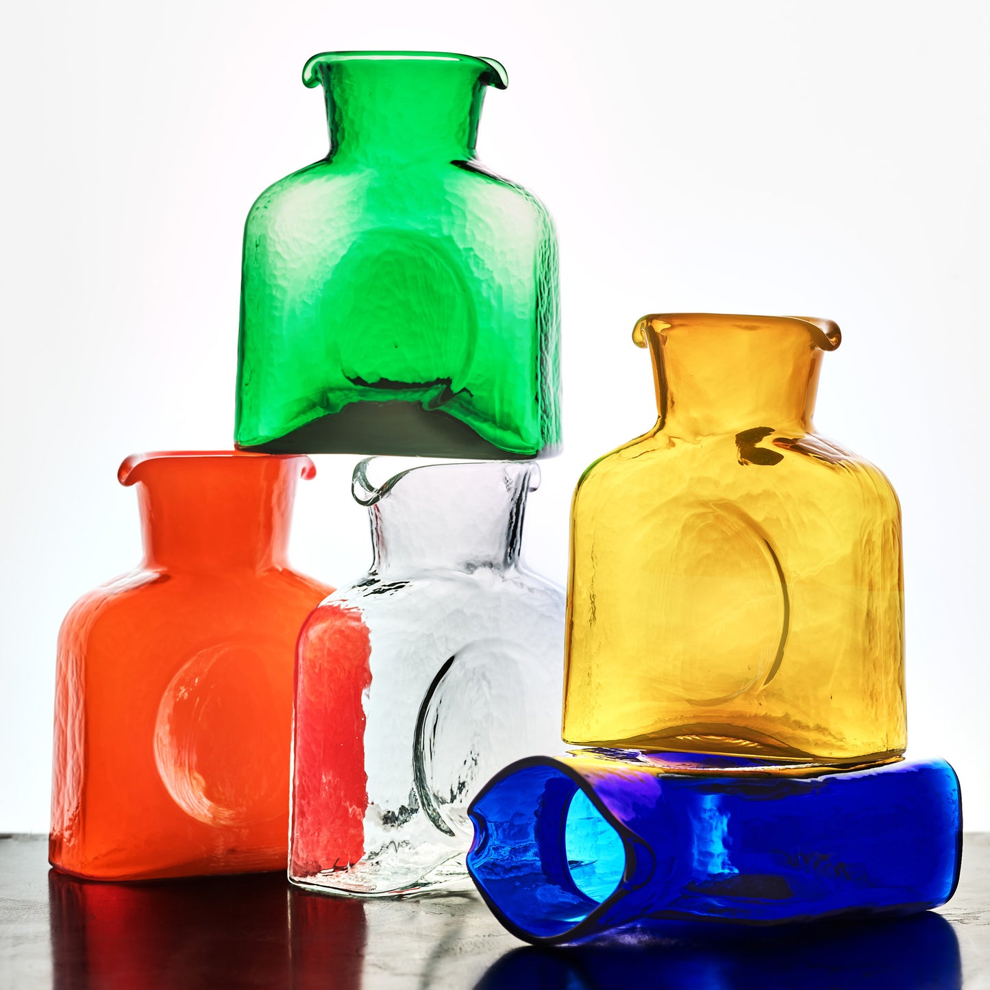 Stack of five colorful square glass pitchers: green, orange, clear, yellow, and blue.