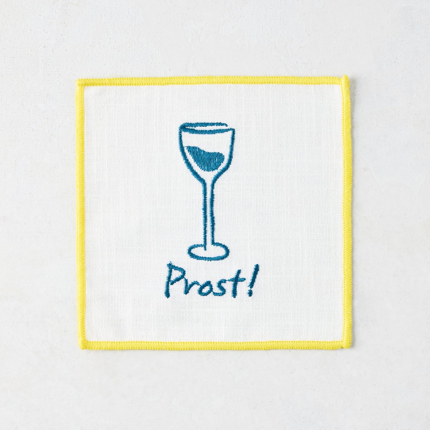 White cotton coaster with yellow trim, blue embroidered wine glass, and "Prost!" text.