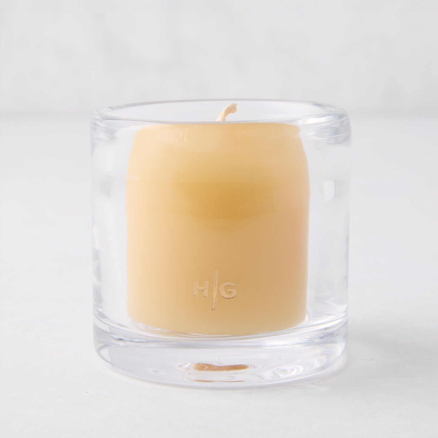 Clear glass votive candle holder with a light yellow candle inside, featuring an "HG" logo.