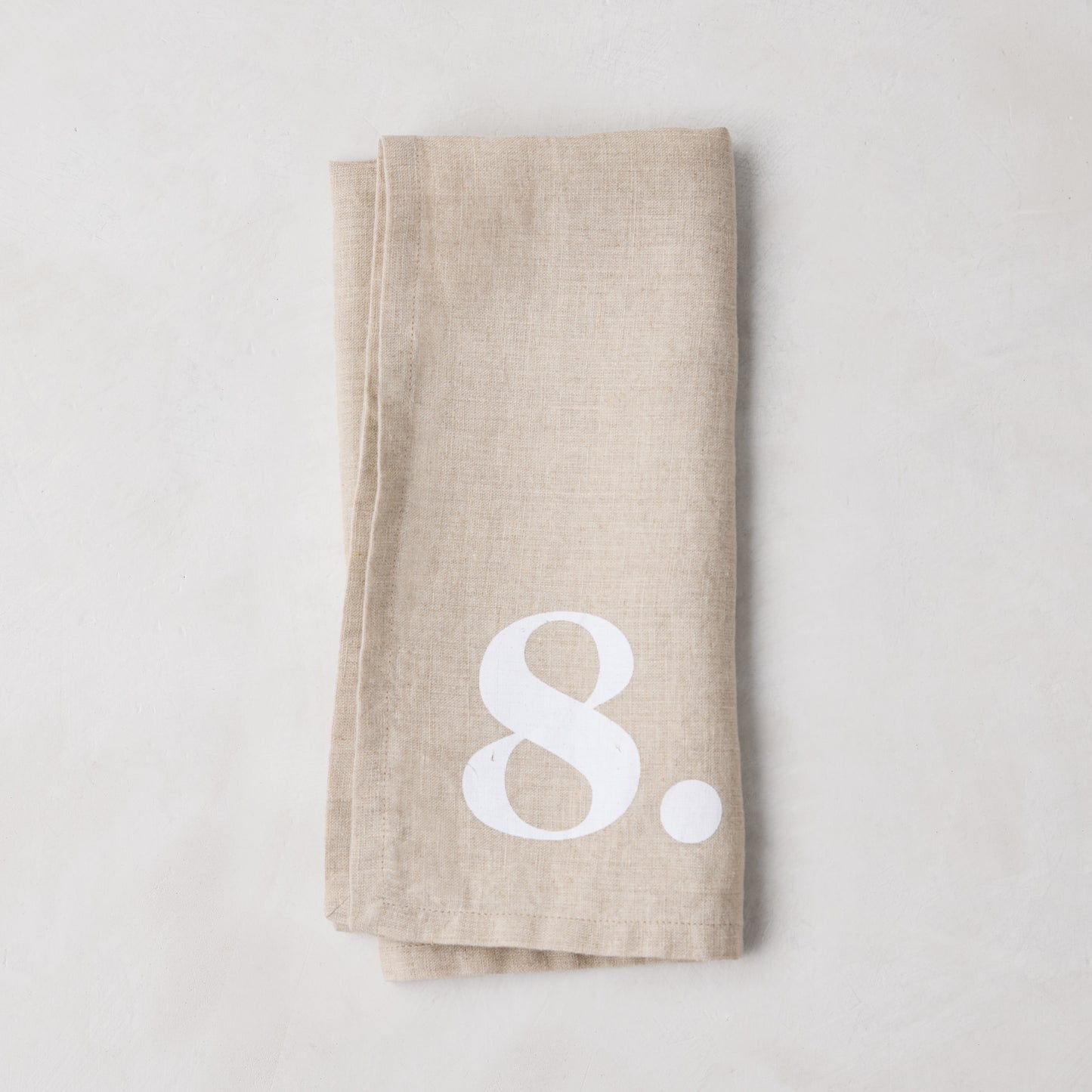 Diane Keaton + Hudson Grace Khaki Linen Numeric Napkin, folded, with a white number 8.