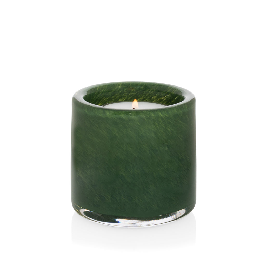 Mouth-blown, hand-formed green votive glass candle holder with a lit candle, reflecting on a white surface.