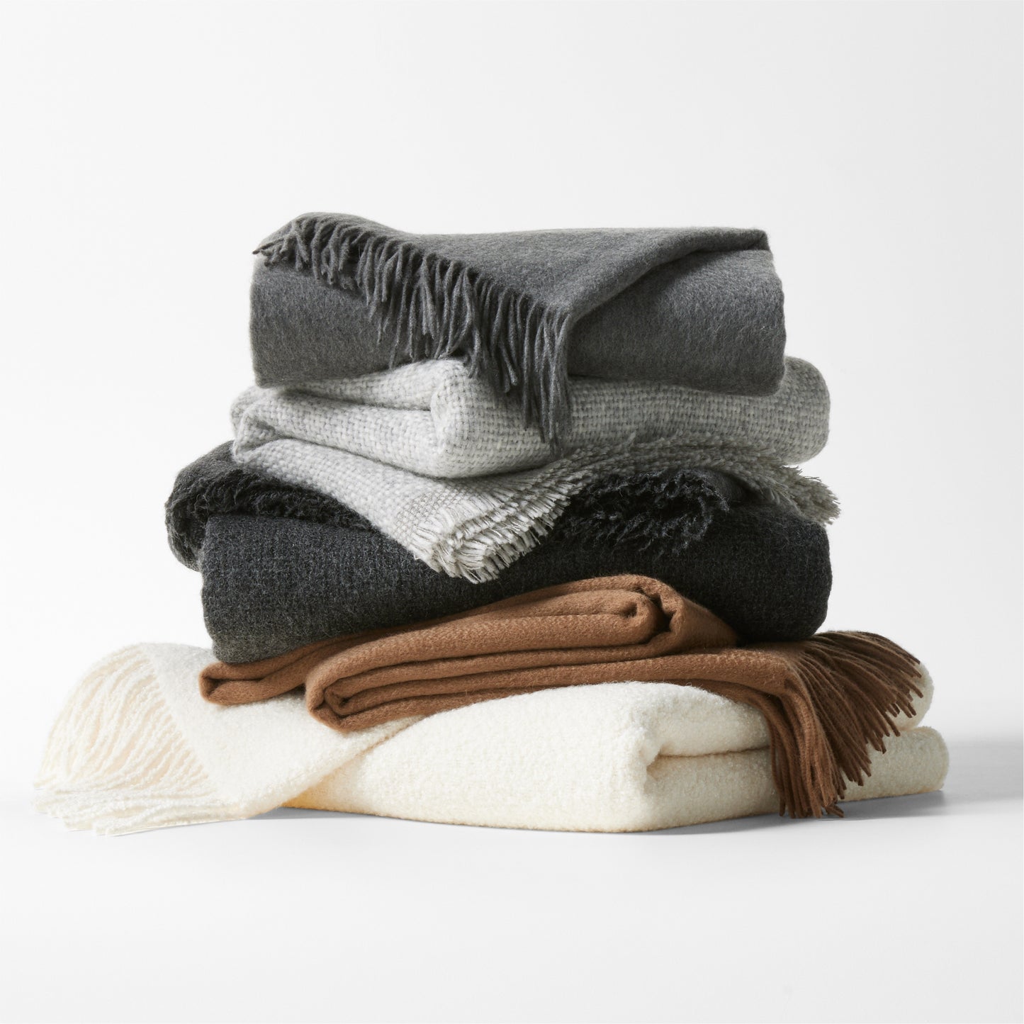 Stack of folded alpaca wool throw blankets in grey, white, cream, and brown with fringed edges.