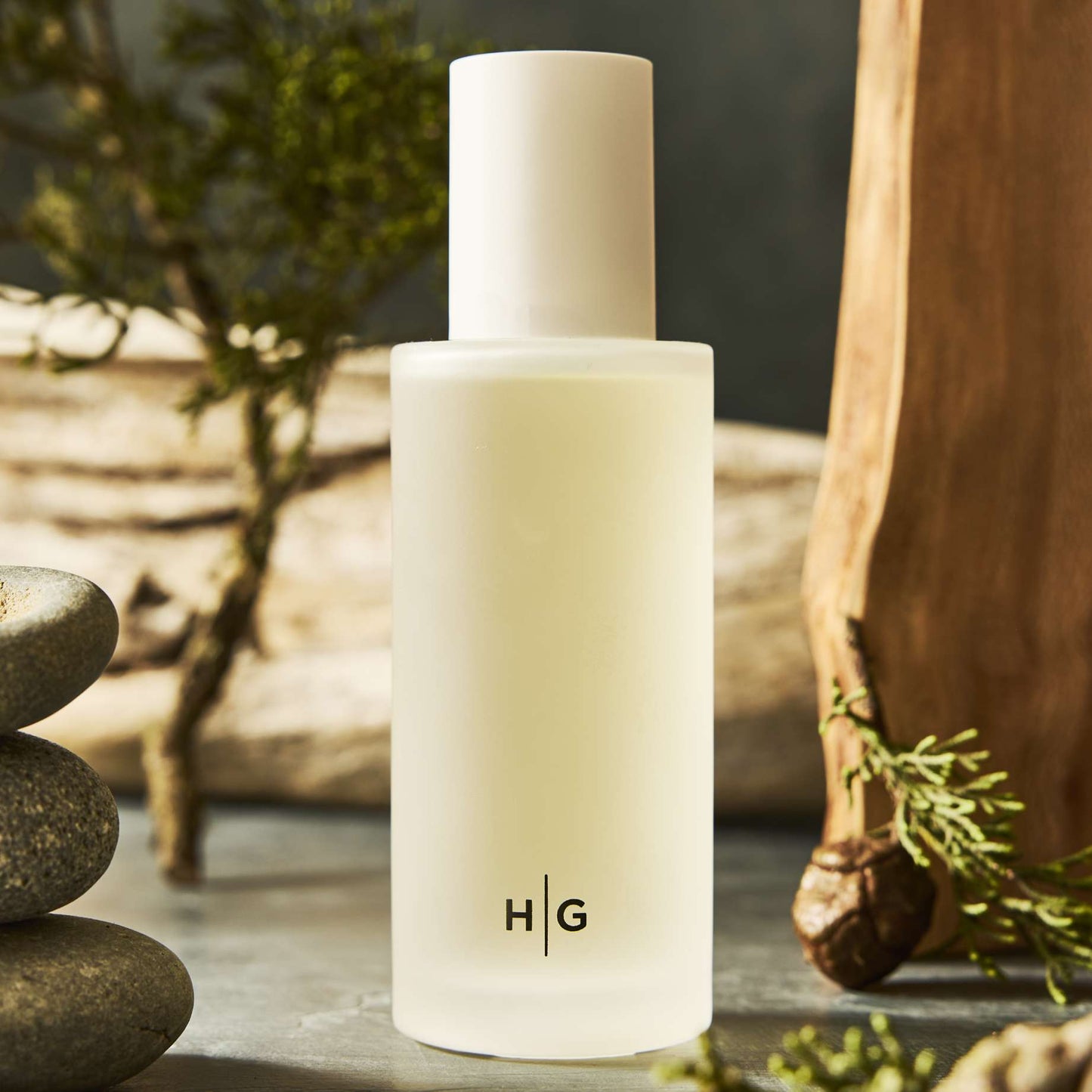 Hudson Grace Brentwood Scented Reed Diffuser: Frosted glass vessel with "H|G" logo, white cap, surrounded by natural wood and greenery.