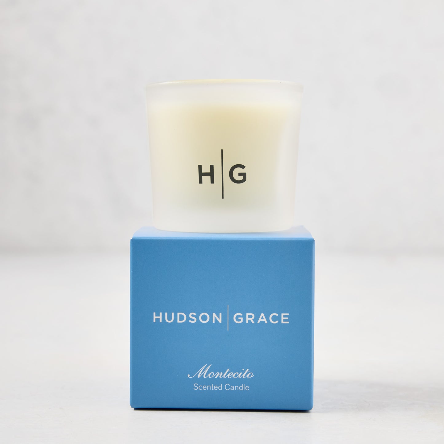 Hudson Grace Montecito scented votive candle in a frosted glass vessel on a blue box.