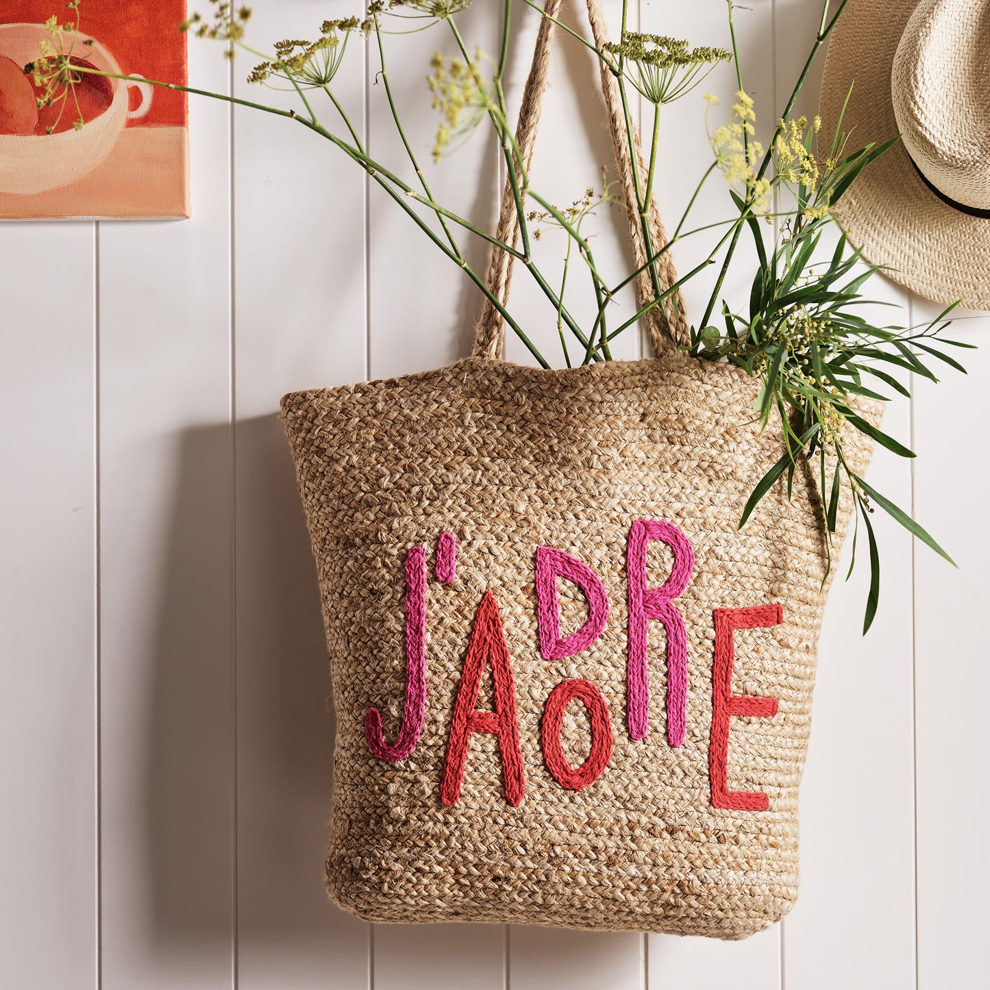 Natural jute woven tote bag with "J'ADORE" embroidered in red and pink, featuring sturdy handles.