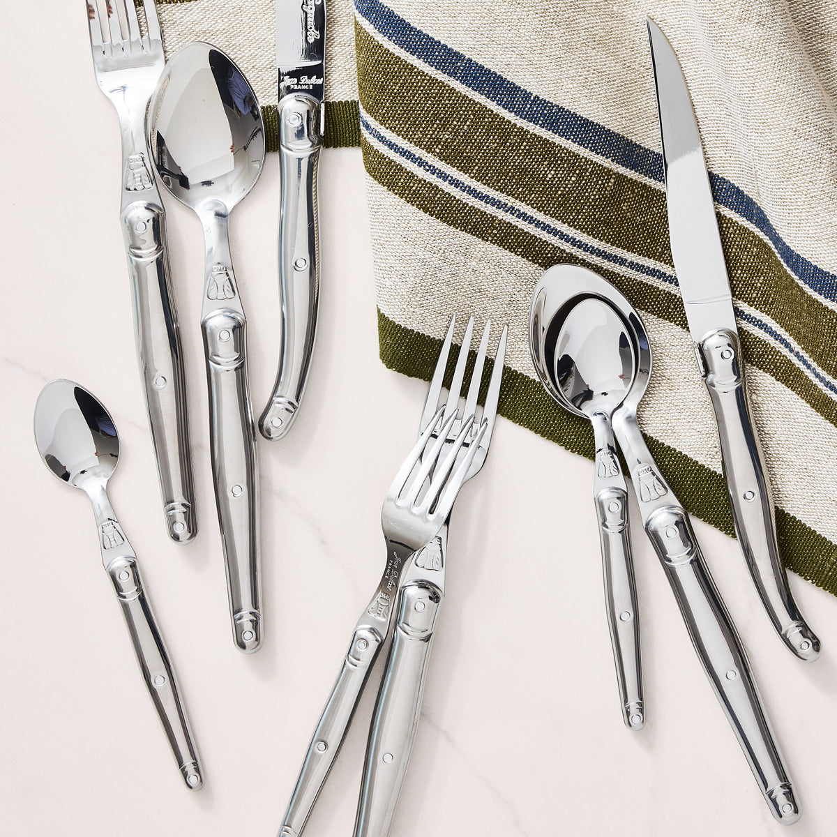 Jean Dubost Laguiole 5-piece stainless steel flatware set with iconic bee emblem on a striped linen napkin.