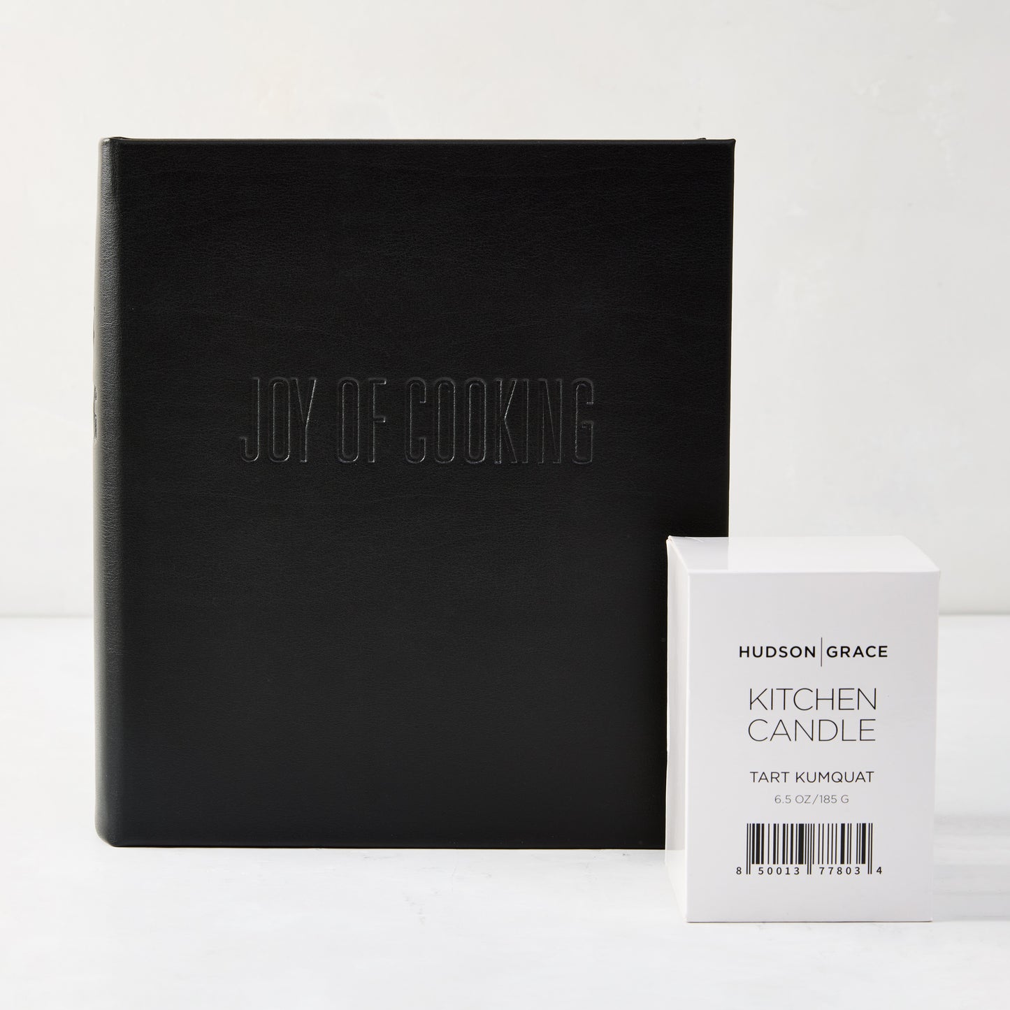 Joy Of Cooking Gift Set: Black leather cookbook with blind embossed title, beside a white Hudson Grace Kitchen Candle box.