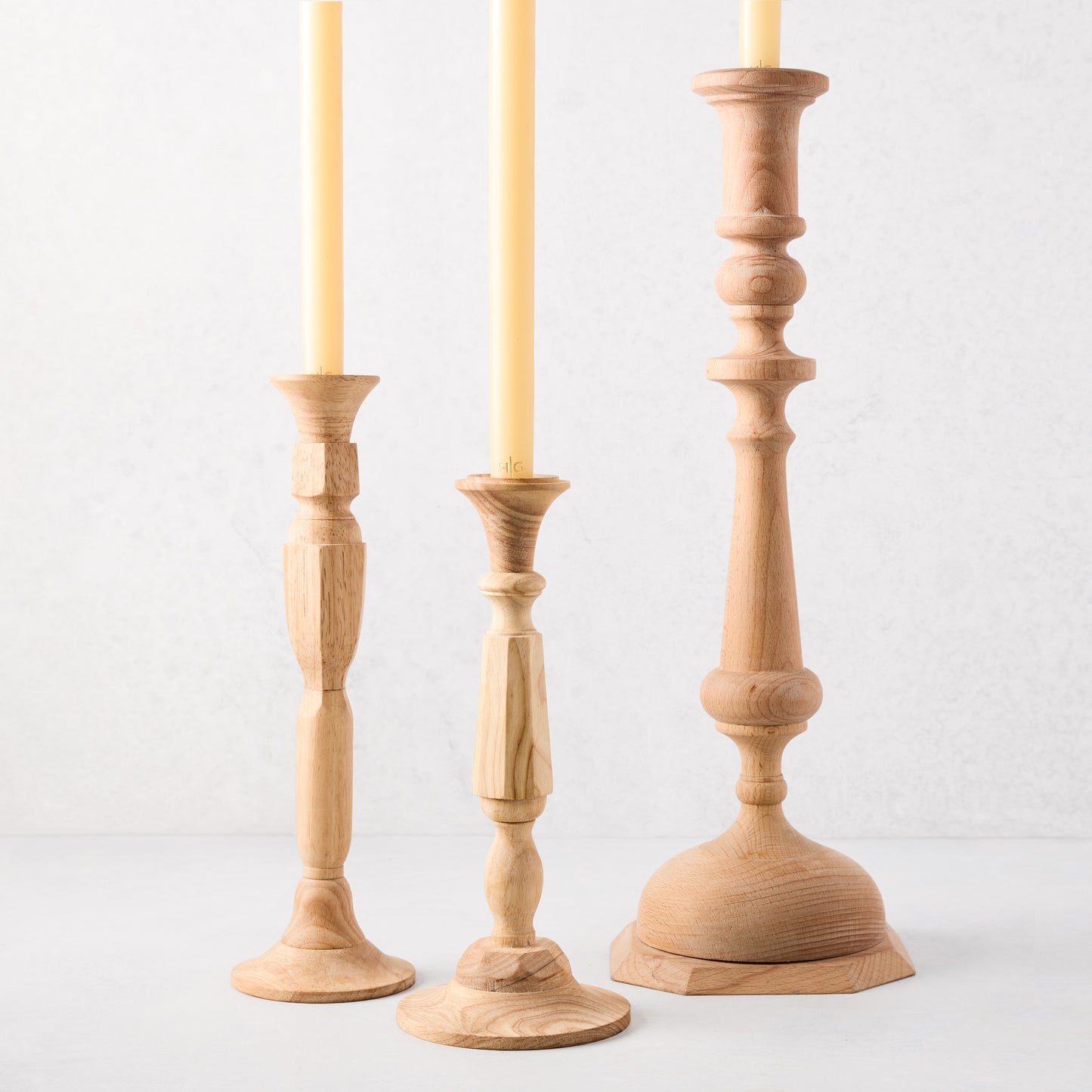 Three natural beechwood Georgian candlesticks of varying heights, holding yellow taper candles.