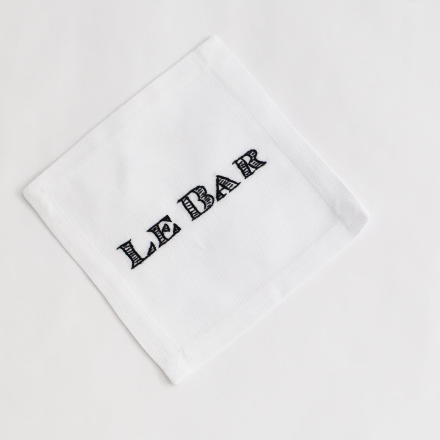 White linen cocktail napkin coaster with "LE BAR" hand-embroidered in black script.