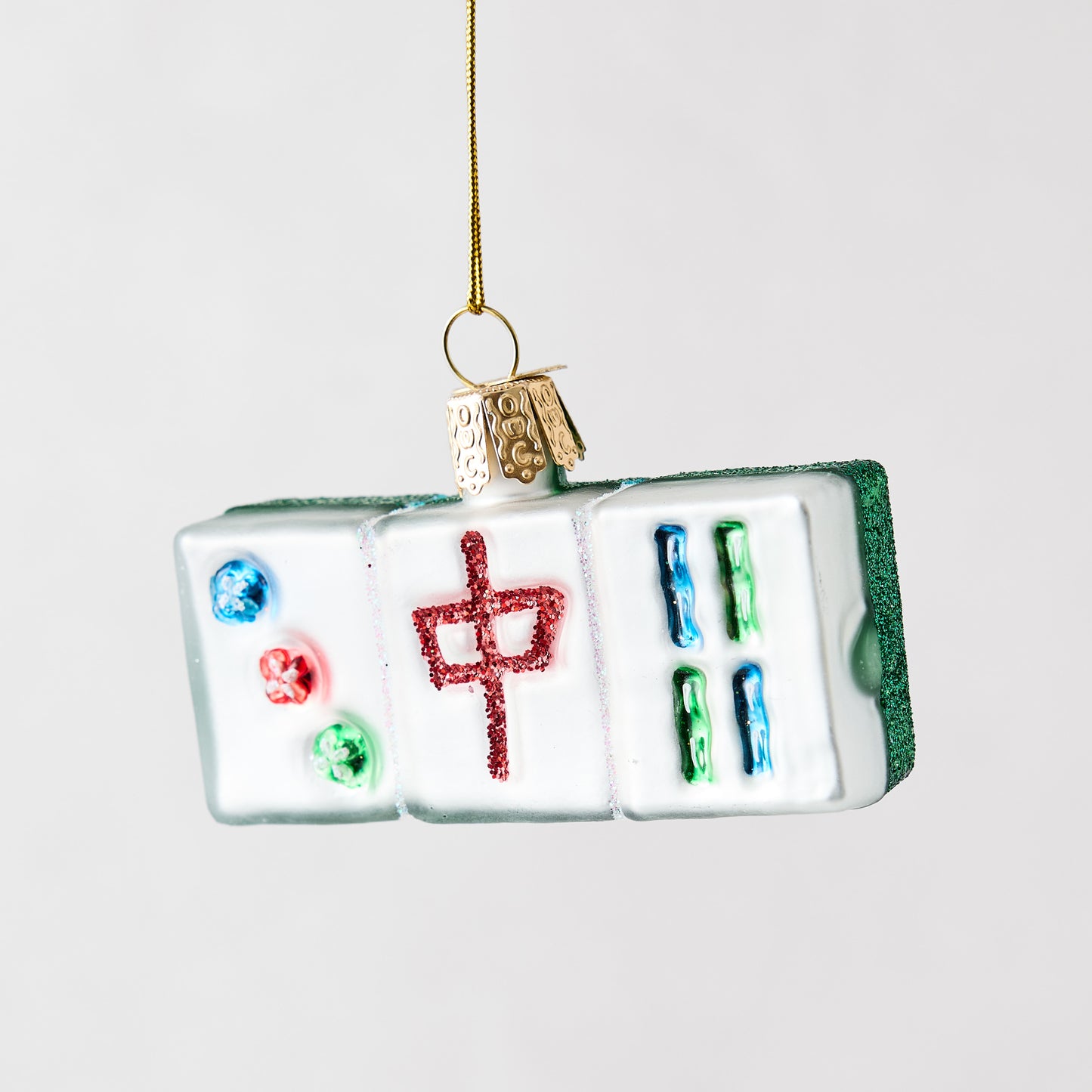 Hand-painted glass Mahjong tile Christmas ornament with glittered red and green details hanging on a gold string.