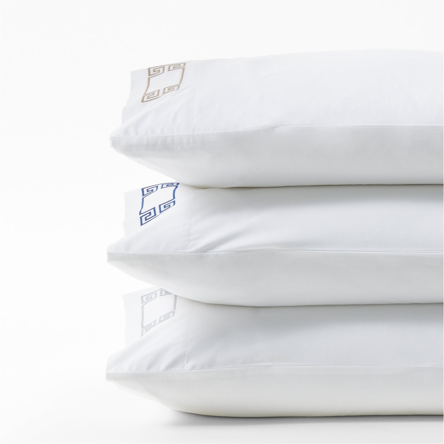 Stack of three white percale pillowcases with embroidered Greek key designs in navy, gold, and silver.