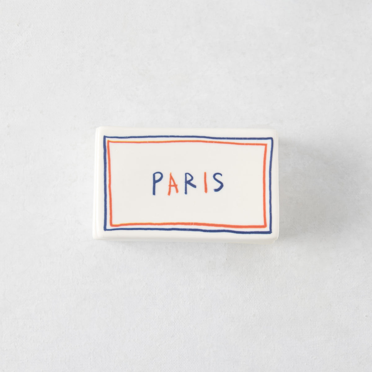 Rectangular white ceramic match box with a hand-painted blue and orange border and the word PARIS in colorful letters.