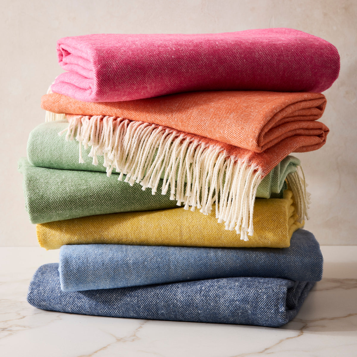 Stacked Italian Herringbone throw blankets in pink, orange, green, yellow, and blue with fringe.