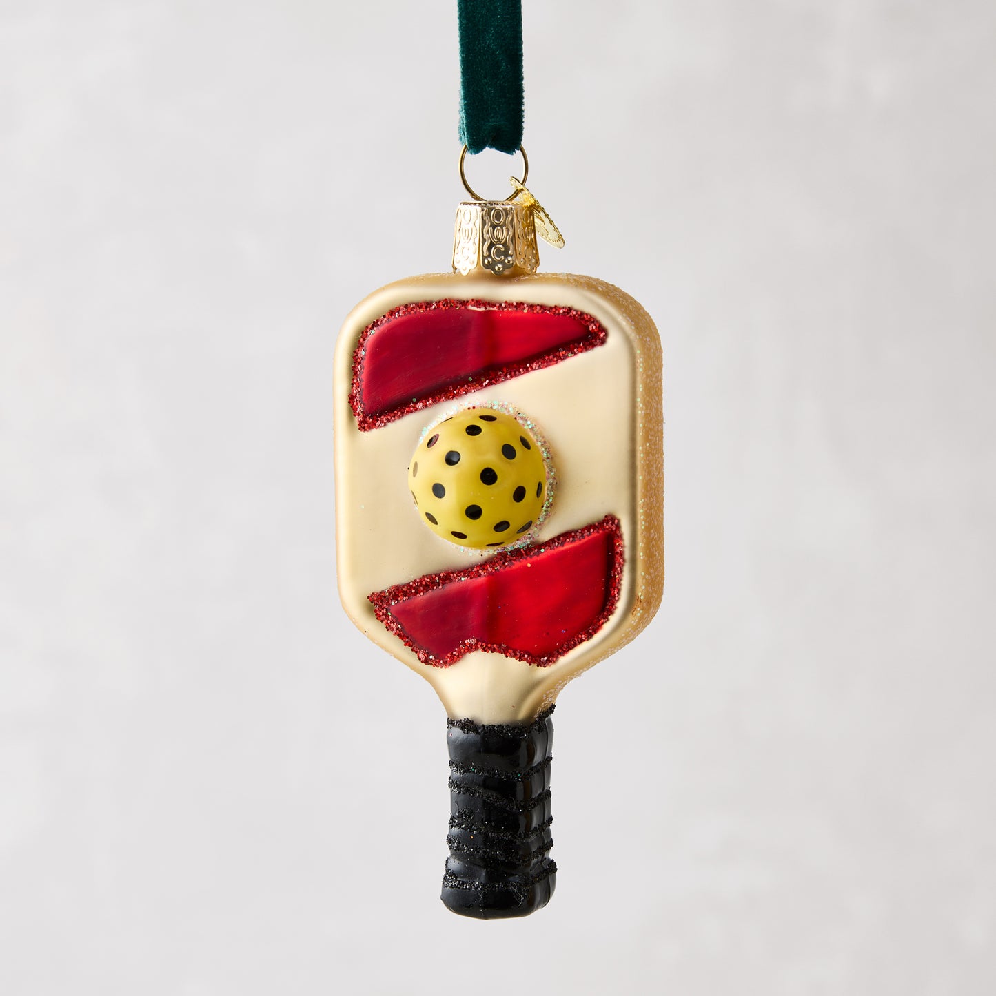 Glittered glass pickleball paddle ornament with red accents, yellow ball, and black handle, hanging from a green ribbon.