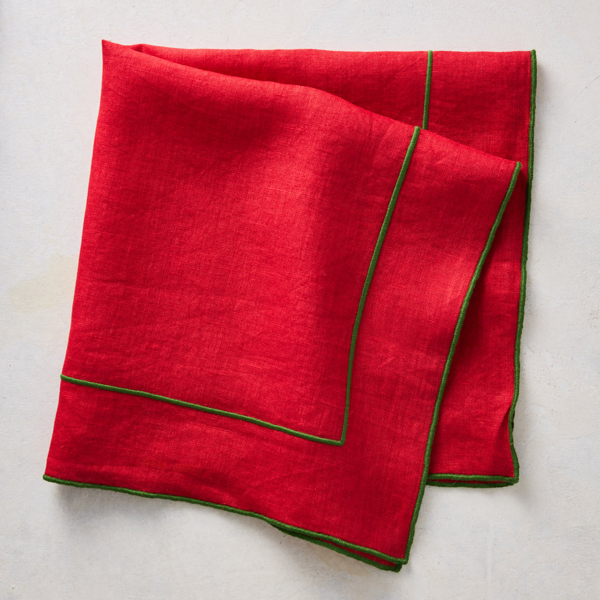Red linen napkin with green embroidered border detail, folded on a neutral textured background.