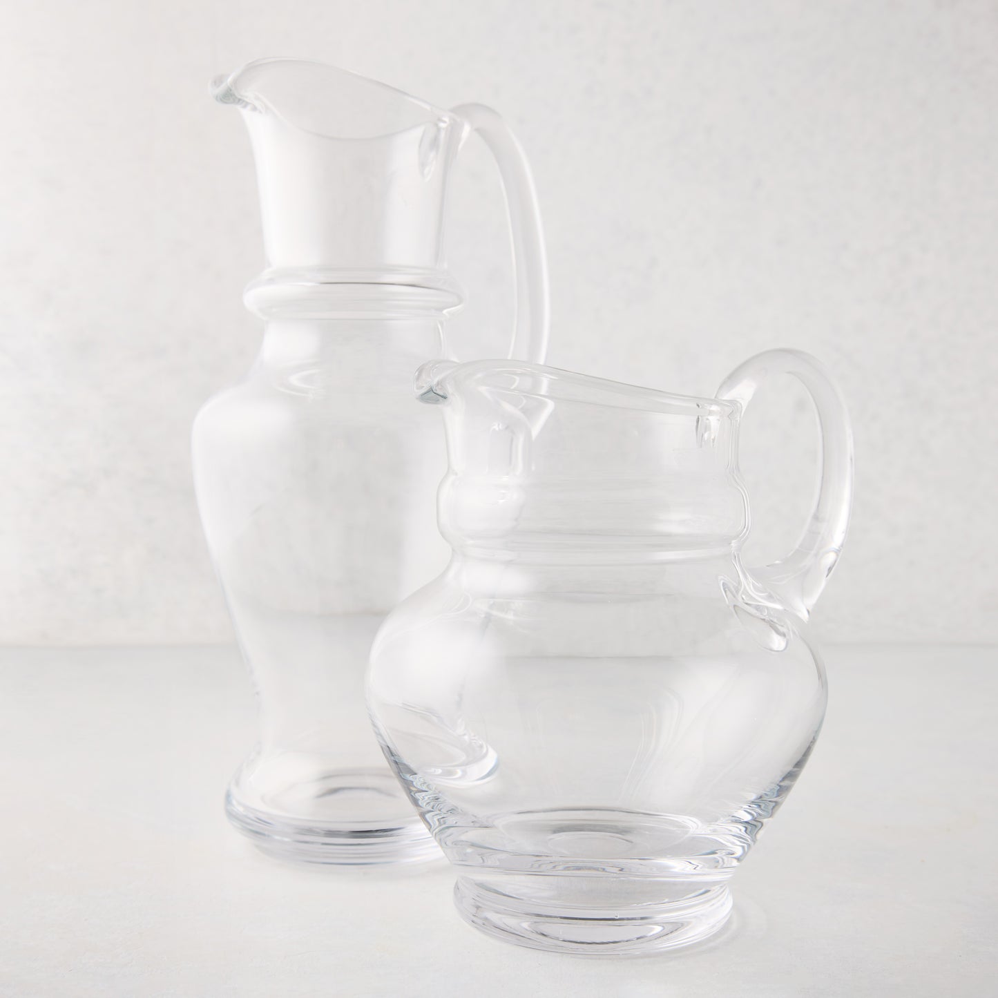 Two clear hand-blown Sutton glass pitchers, one tall and one short, with unique shapes and handles.