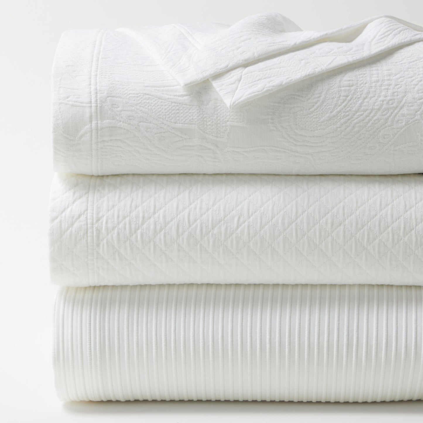 Stacked white cotton matelassé coverlets with diamond, paisley, and ribbed textures.