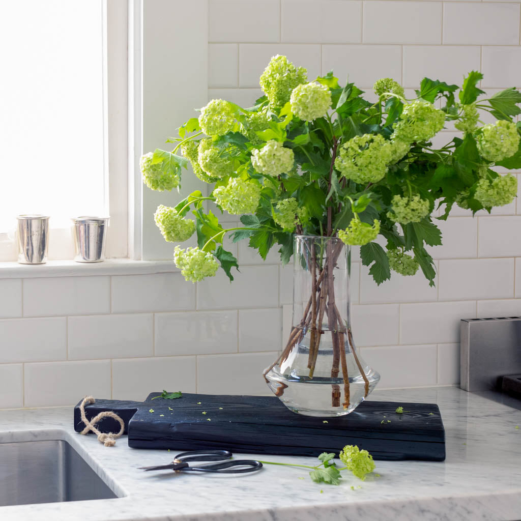 Small black reclaimed wood brasserie serving plank with rope detail, next to a vase of green flowers.