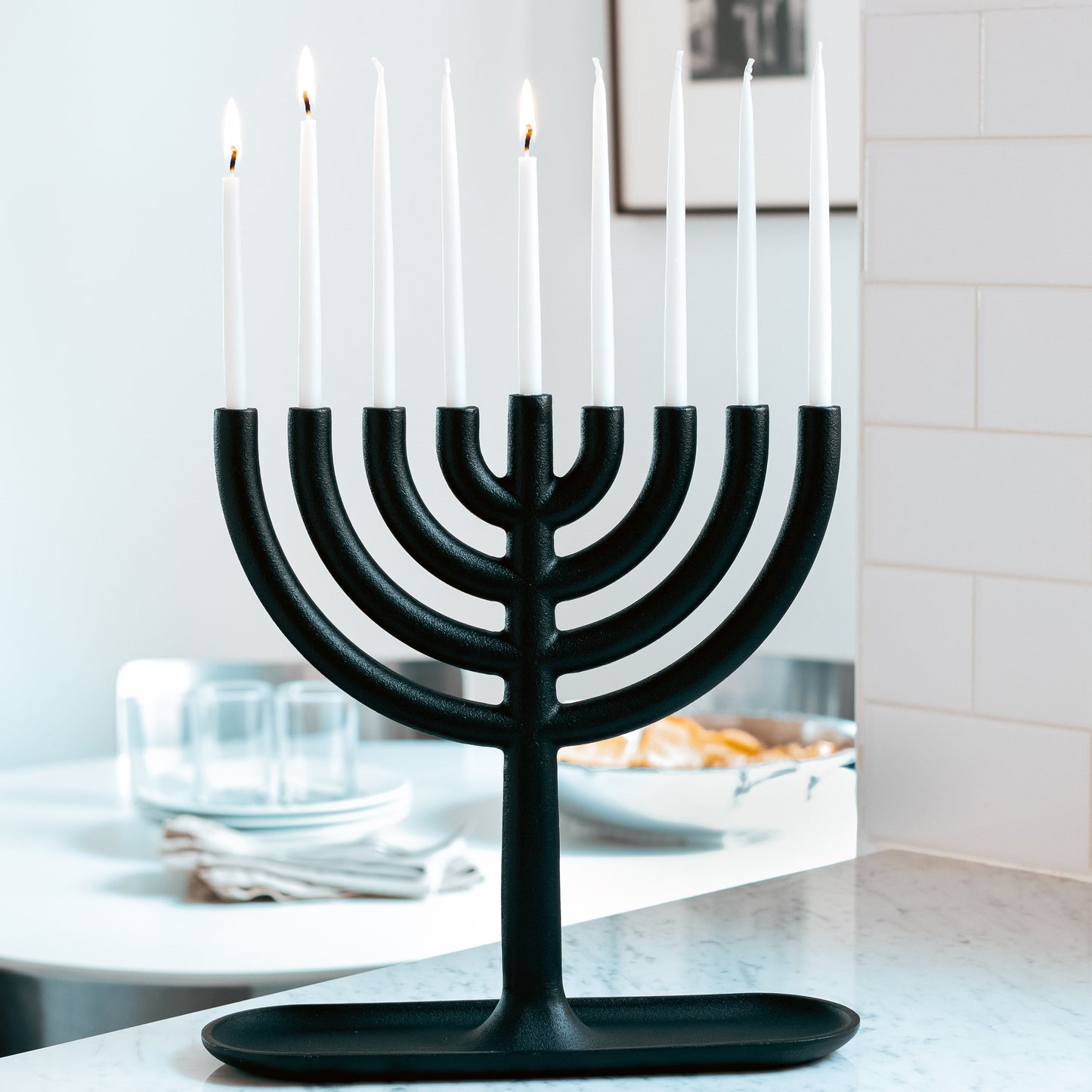 Black cast-iron Menorah with four lit and five unlit white taper candles on a marble counter.