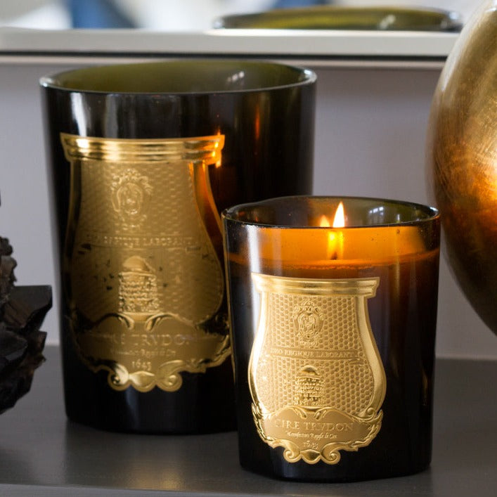 Two Trudon Cyrnos Intermezzo scented candles in dark green glass vessels with gold embossed labels, one lit.