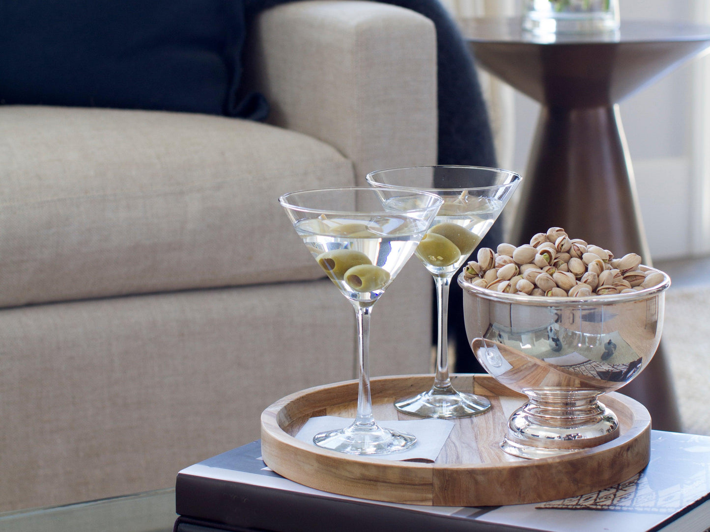 Vintage silverplate 8" footed bowl filled with pistachios, next to two martinis on a wooden tray.