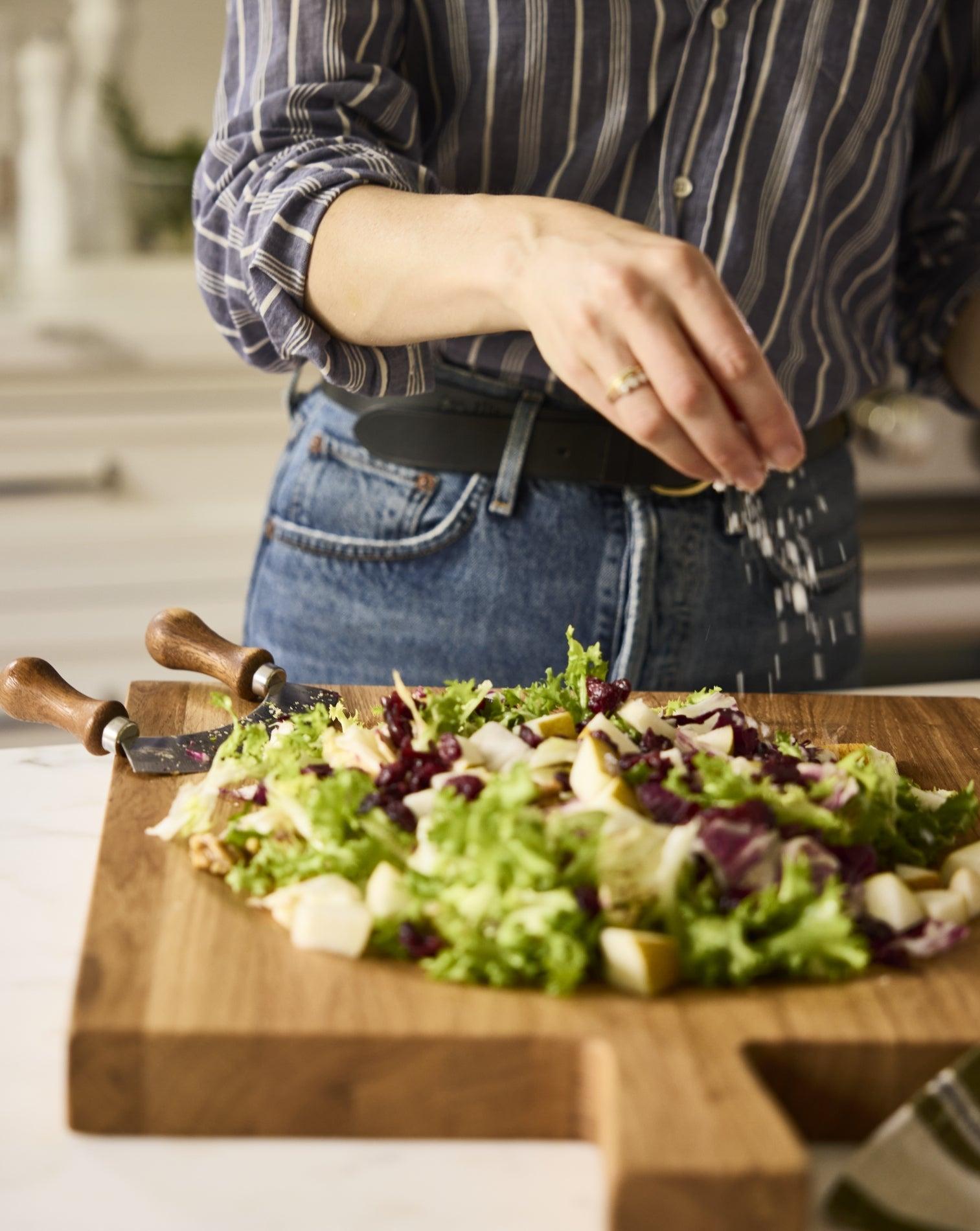 Woman sprinkling cheese on a fresh Thanksgiving salad with cranberries, pears, and greens on a wooden board.