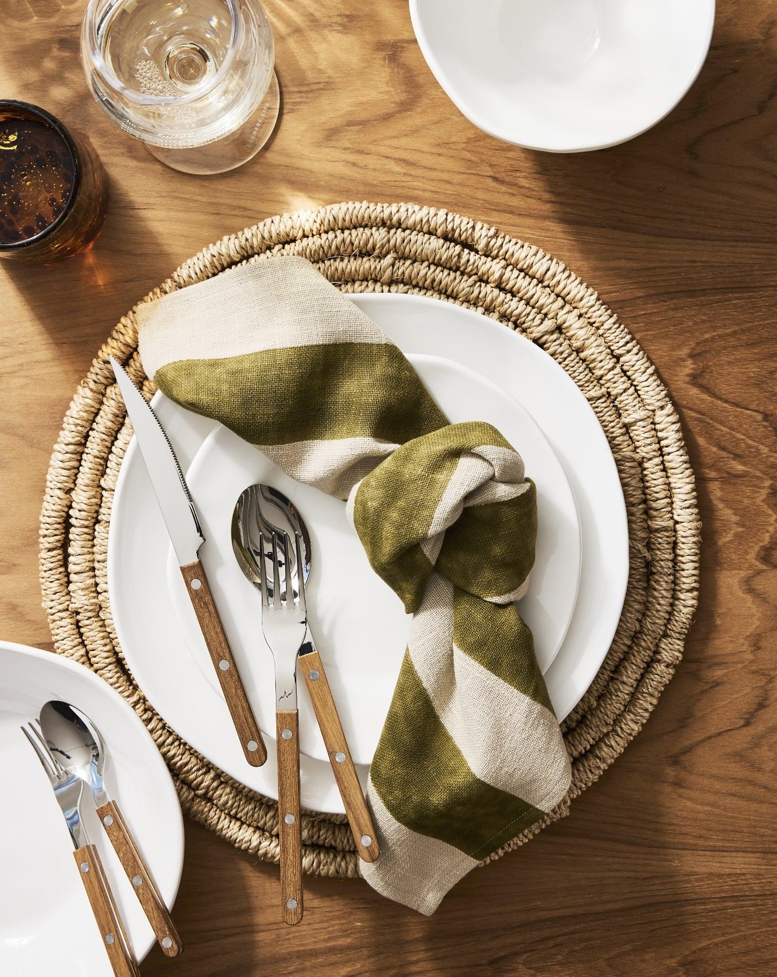Thanksgiving table setting with woven placemat, white plates, wood-handle flatware, and olive striped napkin.