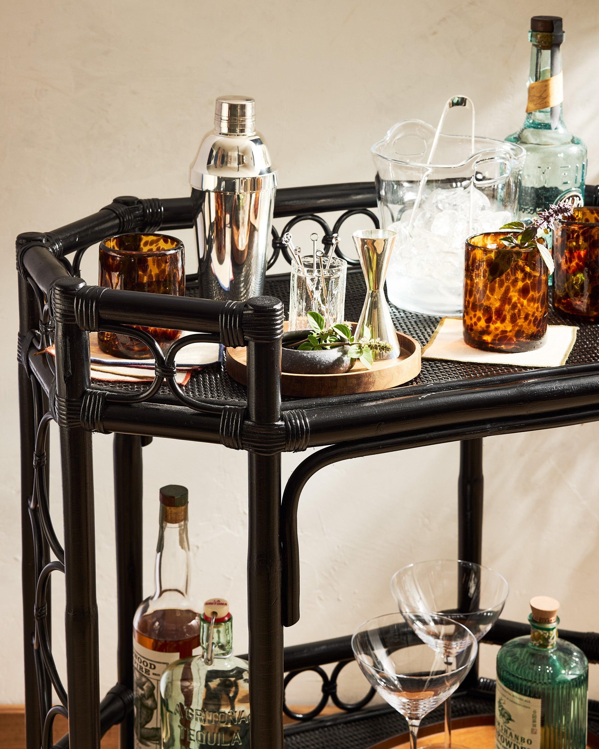 Black rattan bar cart with amber glassware, cocktail shaker, ice bucket, and liquor bottles for The Fall Bar.