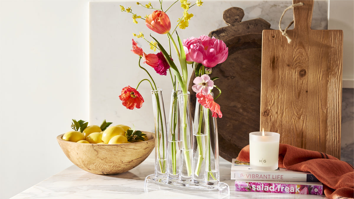 Spring floral arrangement in a glass bud vase next to a wooden bowl of lemons and a lit candle on a marble countertop.