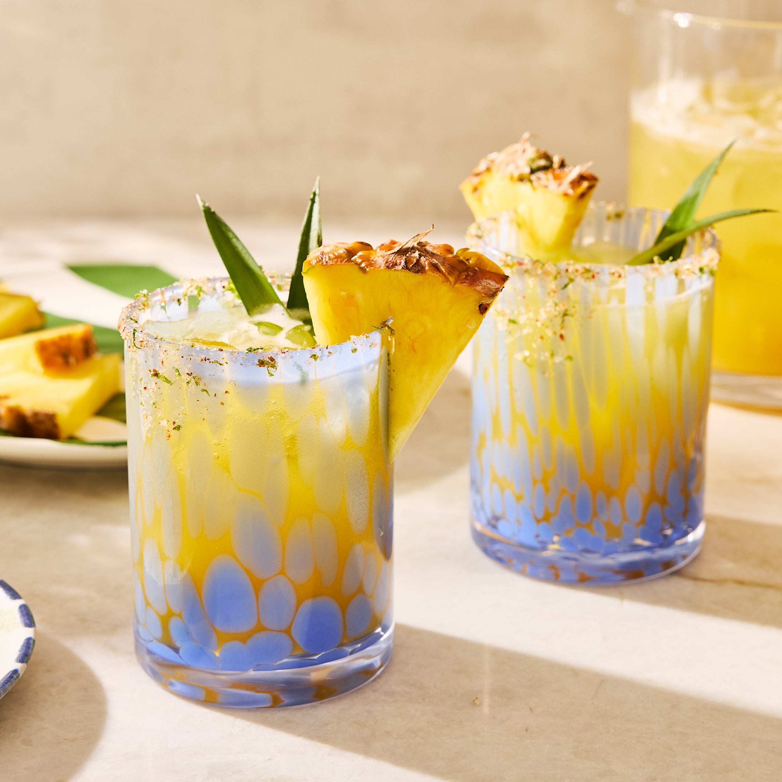 Two pineapple margaritas in blue and yellow patterned glasses, garnished with fresh pineapple wedges and green leaves.