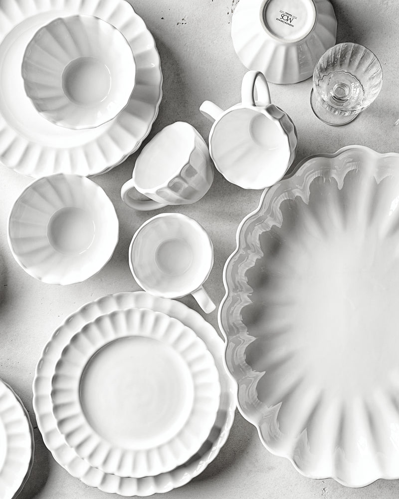 White ceramic dinnerware with scalloped edges arranged on a light gray surface, including plates, bowls, and mugs.