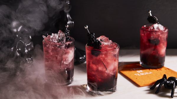 Two red blackberry cocktails with ice and metal picks on an orange Trick or Tequila napkin amidst spooky dry ice smoke.