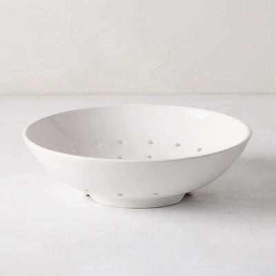 A white ceramic berry bowl with small holes for drainage, placed on a light surface.