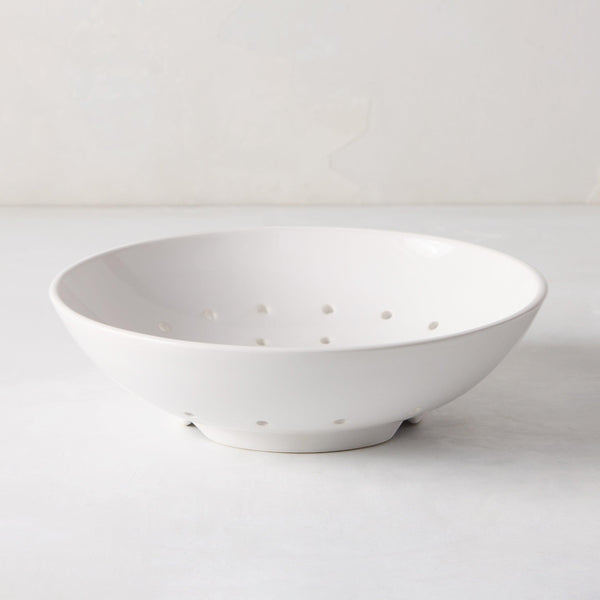 A white ceramic berry bowl with small holes for drainage, placed on a light surface.