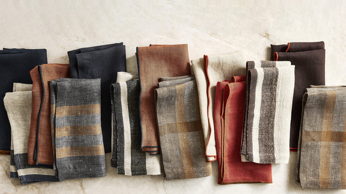 A collection of folded linen napkins in various colors and patterns, including plaid and stripes, arranged neatly on a light beige marble surface.