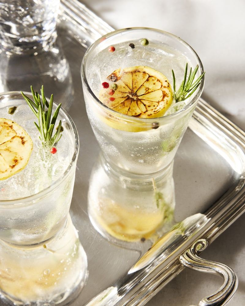 Two gin and tonics garnished with charred lemon slices, rosemary sprigs, and peppercorns on a silver tray.