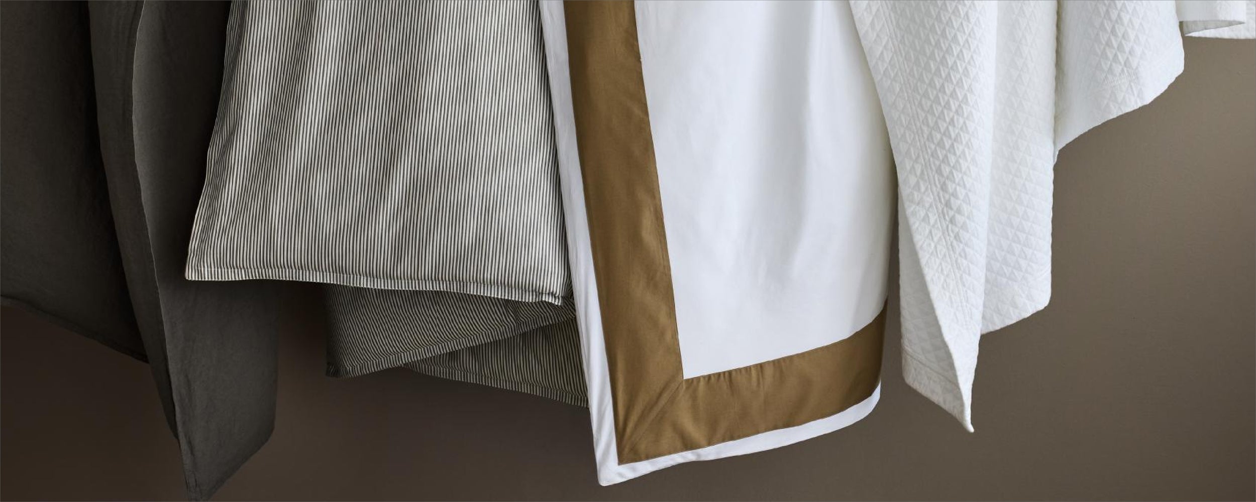 Layered bedding featuring a grey striped sheet, a white duvet with a brown border, and a white quilted coverlet.