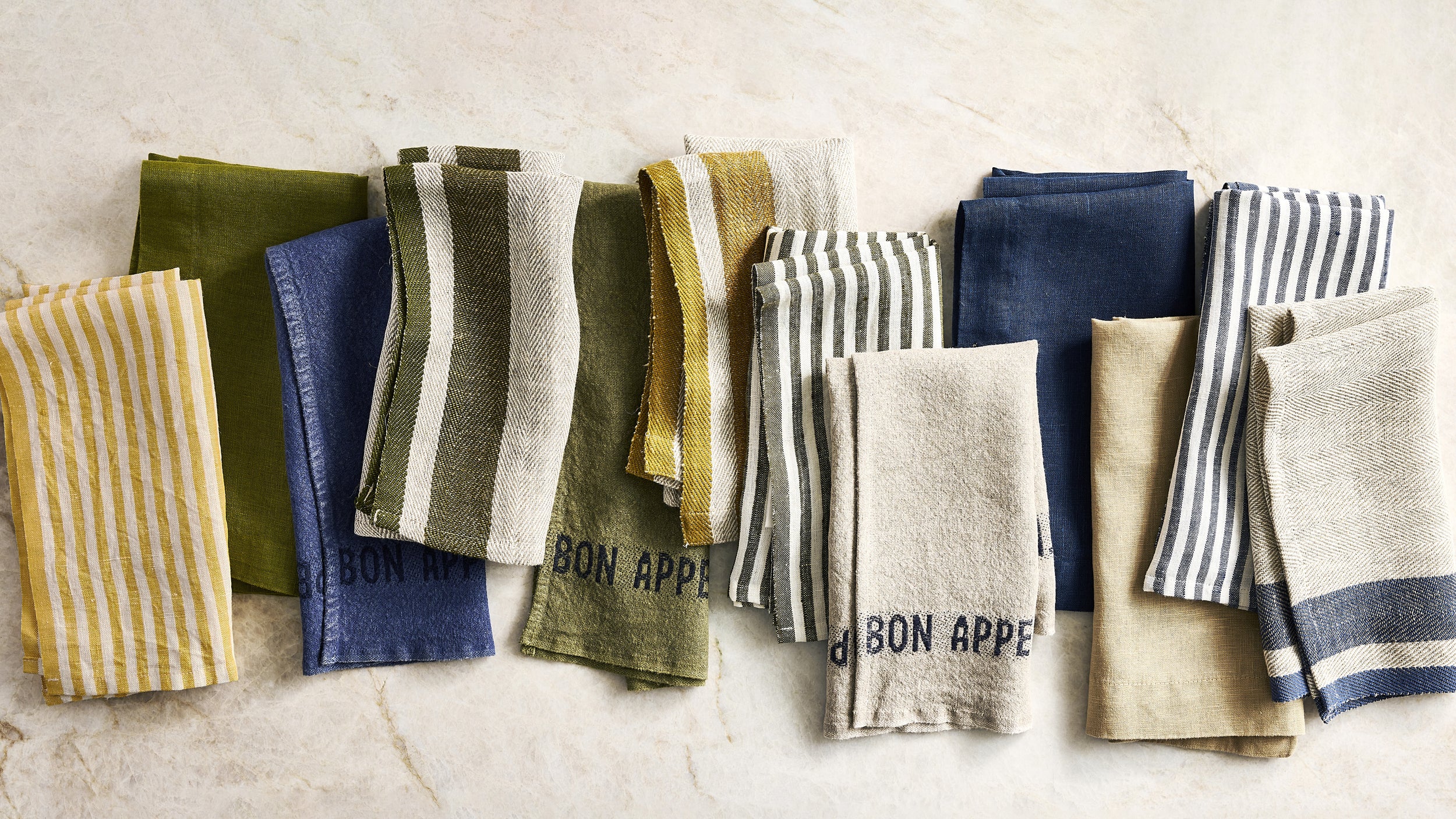 A set of folded striped and solid cotton kitchen towels in various colors including yellow, blue, olive green, beige, and navy, arranged on a light beige marble surface. Some towels feature the text 'BON APPETIT'.