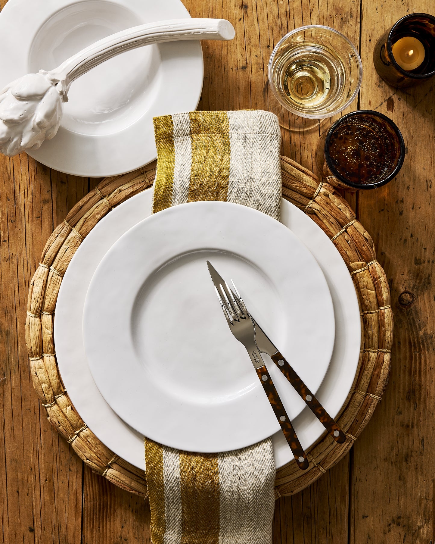 A styled tabletop setting on a rustic wooden table featuring a white ceramic dinner plate layered over a woven round placemat, with a striped cream and mustard napkin, a fork and knife with dark wood handles, a white bowl, a glass of white wine, and two dark candles.