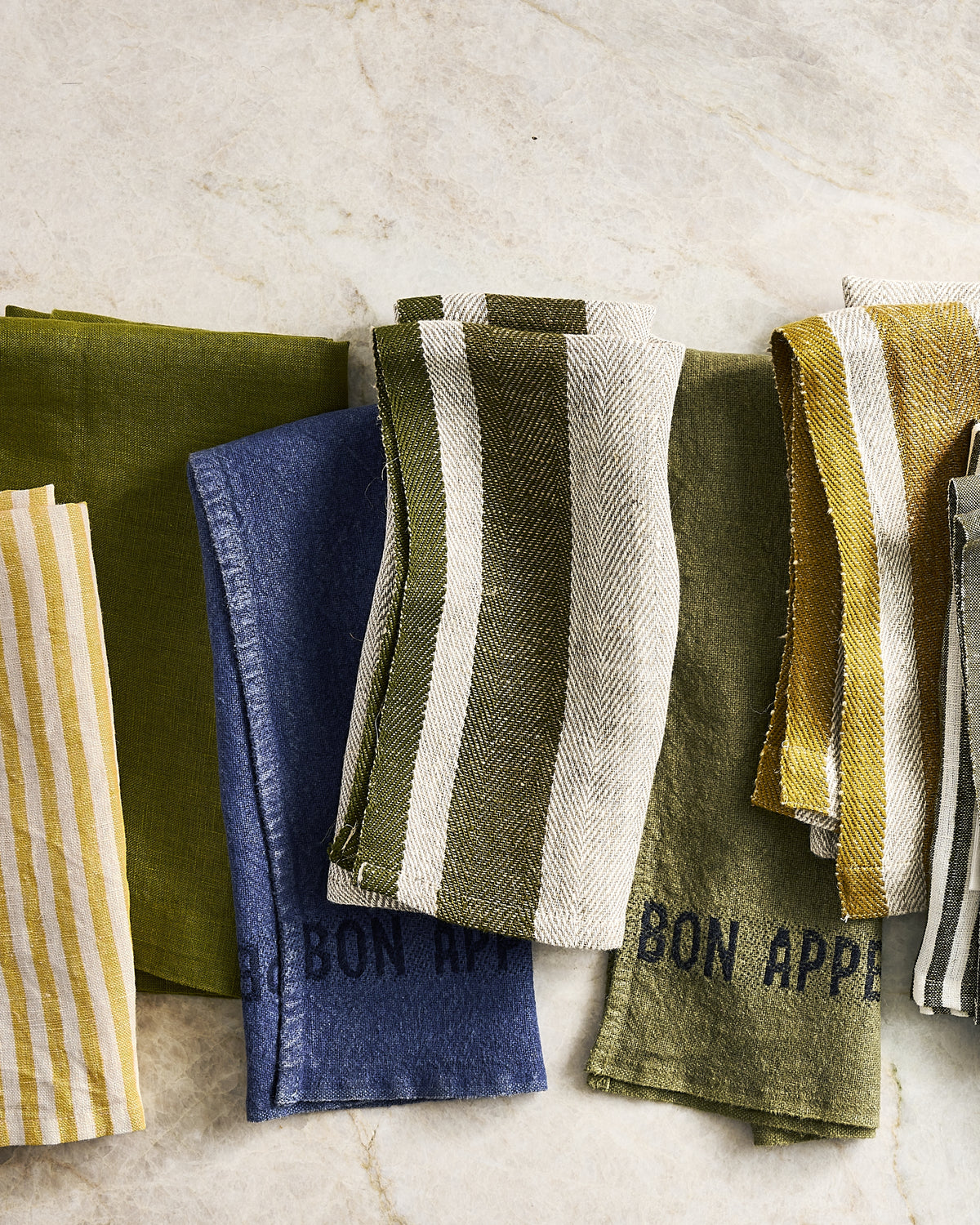 A set of folded linen kitchen towels in various patterns, including solid olive green, blue, yellow stripes, and green-and-white stripes, laid out on a light beige marble surface. Some towels feature the text BON APPETIT