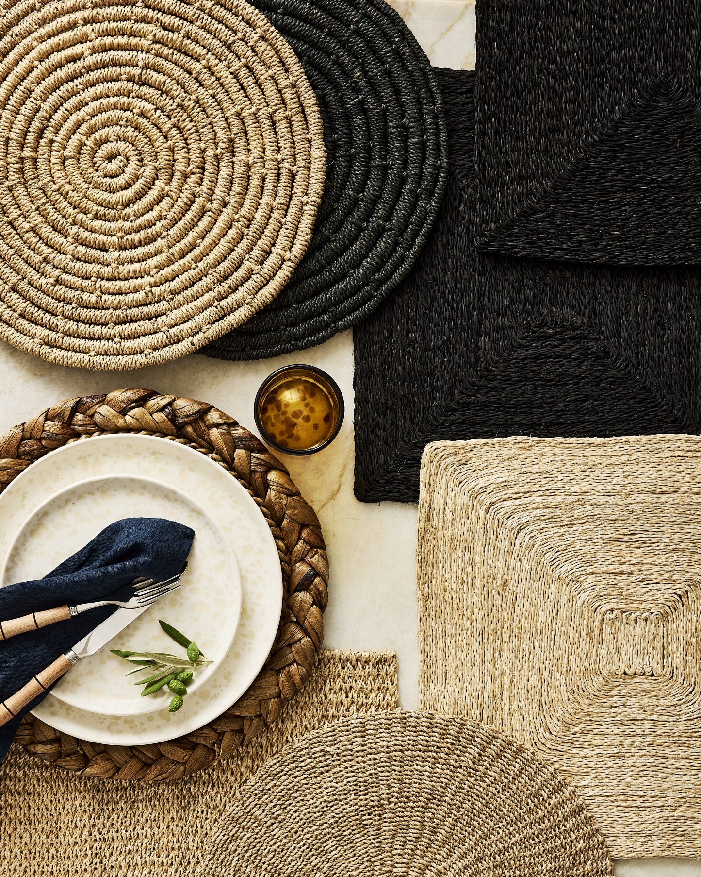 A styled flat-lay of woven placemats and table textiles in natural beige, tan, and black, arranged around white plates, a navy napkin, cutlery, and a small amber glass candle on a light surface.