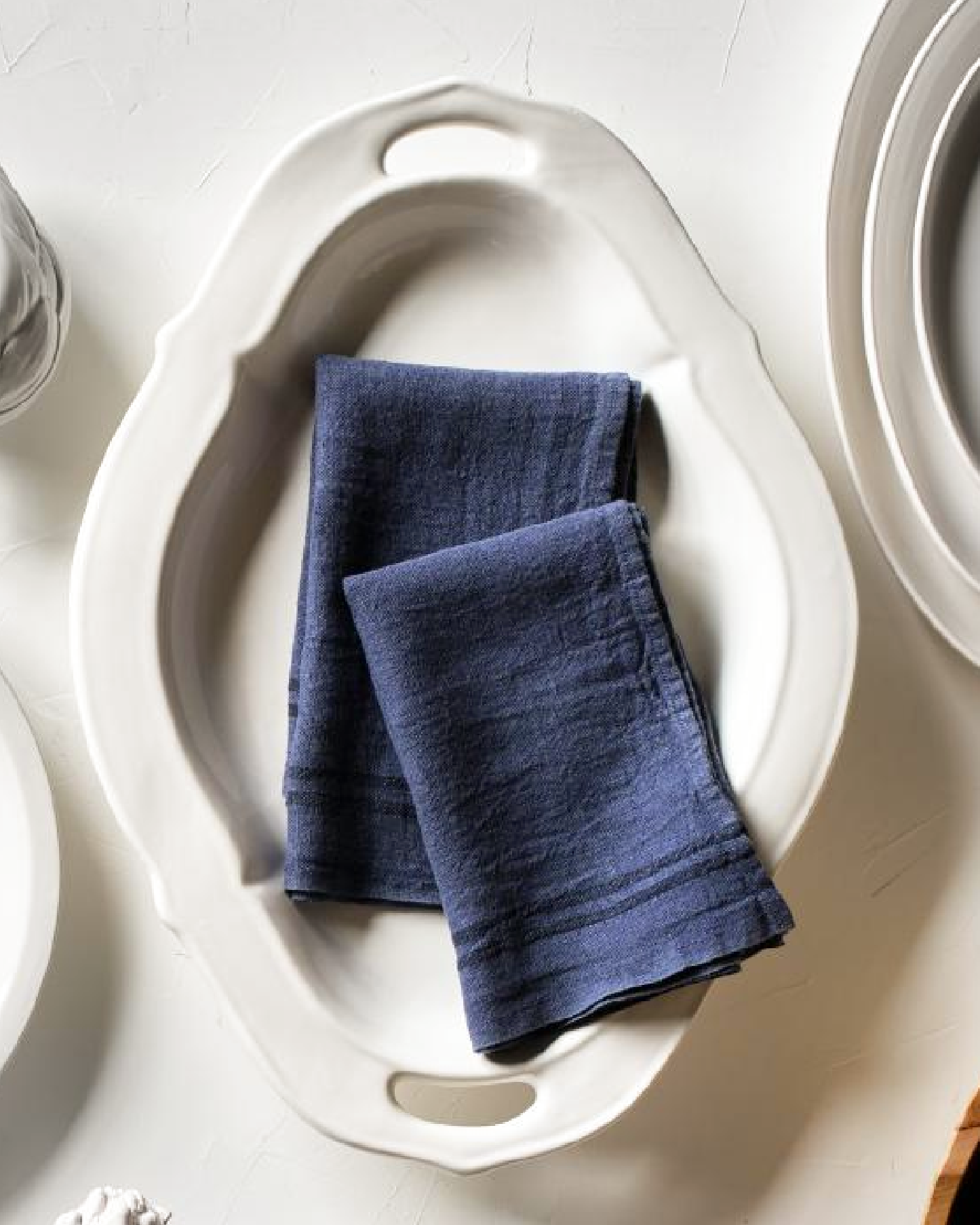 Two folded navy blue linen napkins placed inside an off-white ceramic serving dish on a light tabletop.
