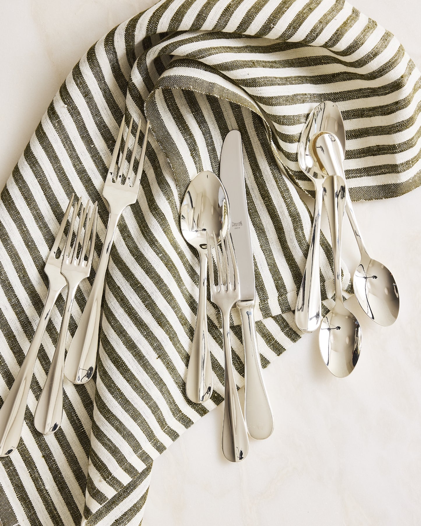 A flat-lay of polished silver flatware, including forks, knives, and spoons, arranged on a cream and olive green striped linen napkin over a light marble surface.