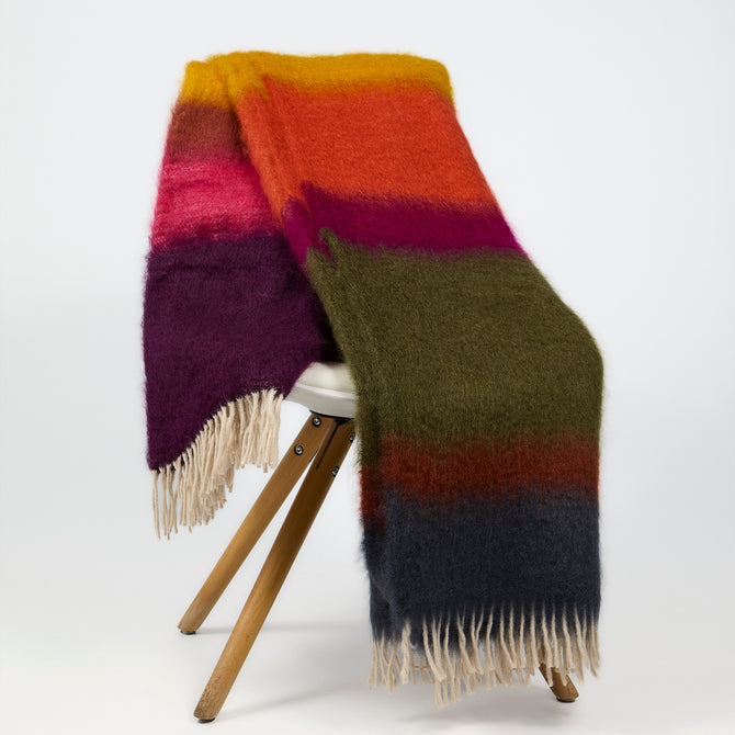 A fuzzy striped wool blanket with cream fringe, draped over a wooden stool against a plain white studio background.