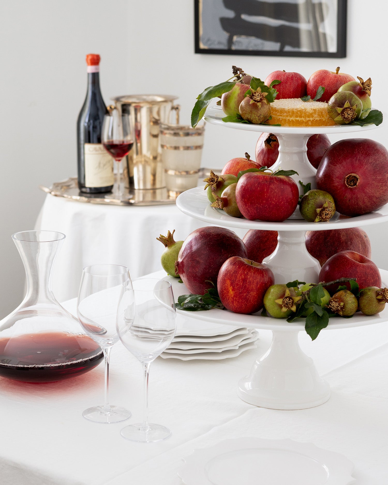 Rosh Hashanah pomegranates on a tiered stand with wine and glasses on a table.