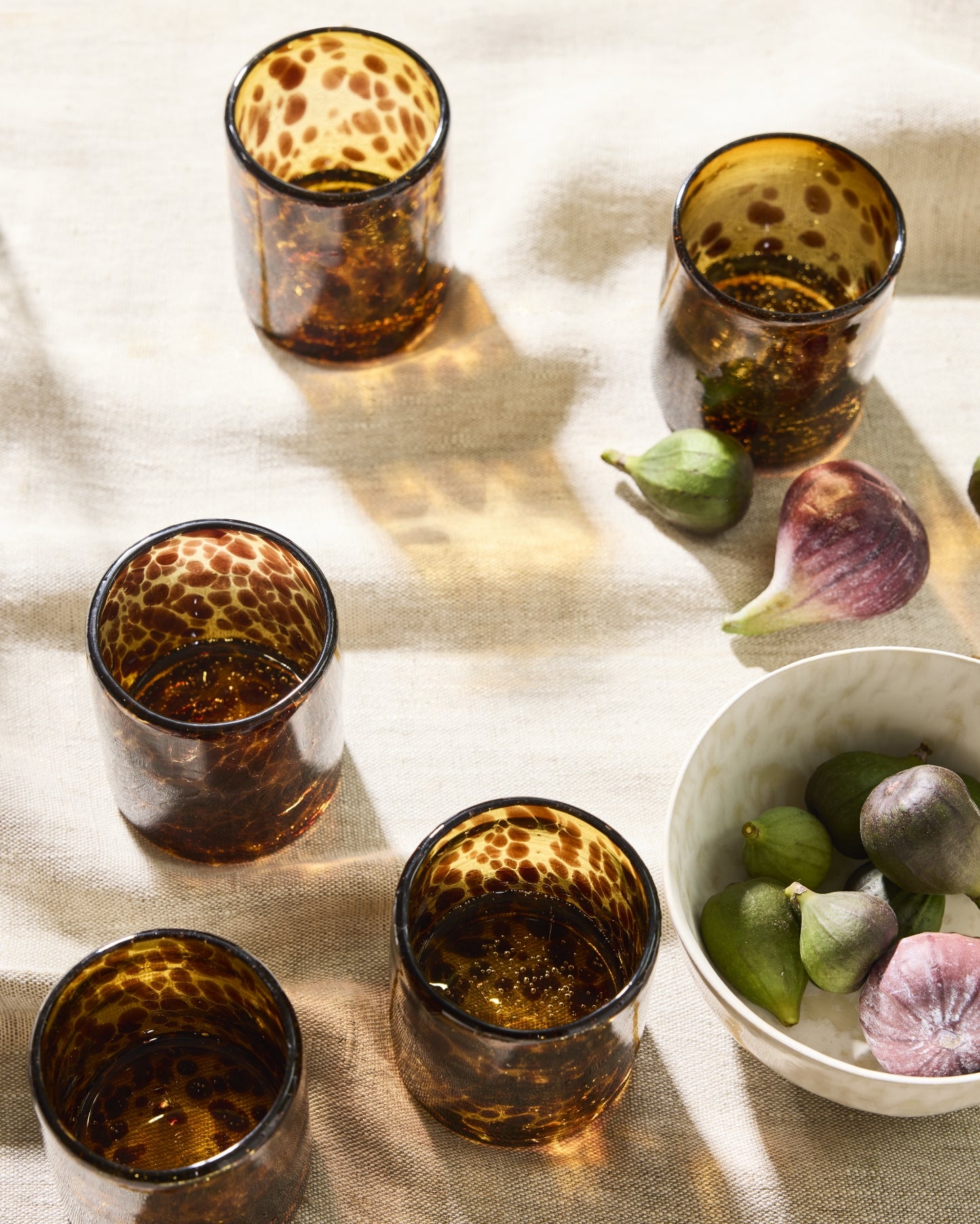 Five amber glass tumblers with a dark spotted pattern, arranged on a beige linen cloth alongside fresh figs and a white bowl.